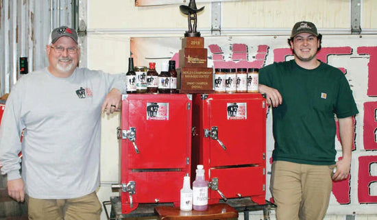 Two BBQ experts proudly display championship trophy and sauces in front of smokers, showcasing top grilling and BBQ skills.