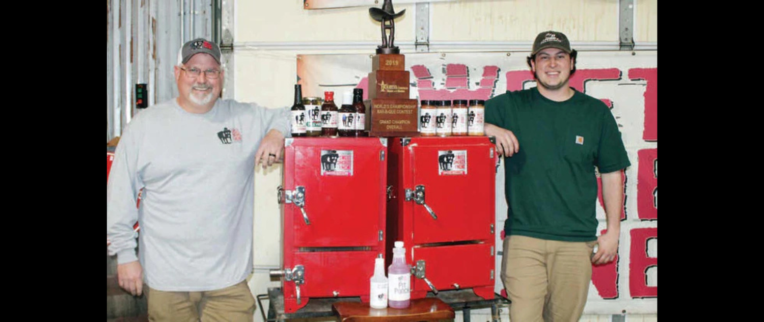 Two BBQ enthusiasts with trophy and BBQ sauces in front of red smokers, showcasing BBQ lifestyle and competition spirit.